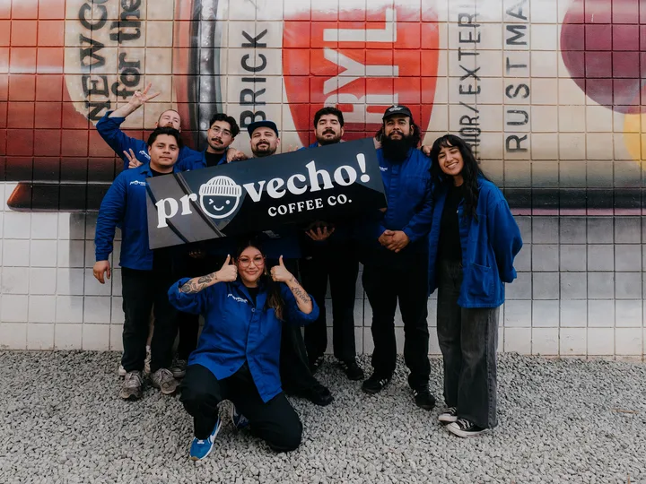 The Provecho team in Provecho workwear holding a Provecho! Coffee Co. banner in front of a painted mural