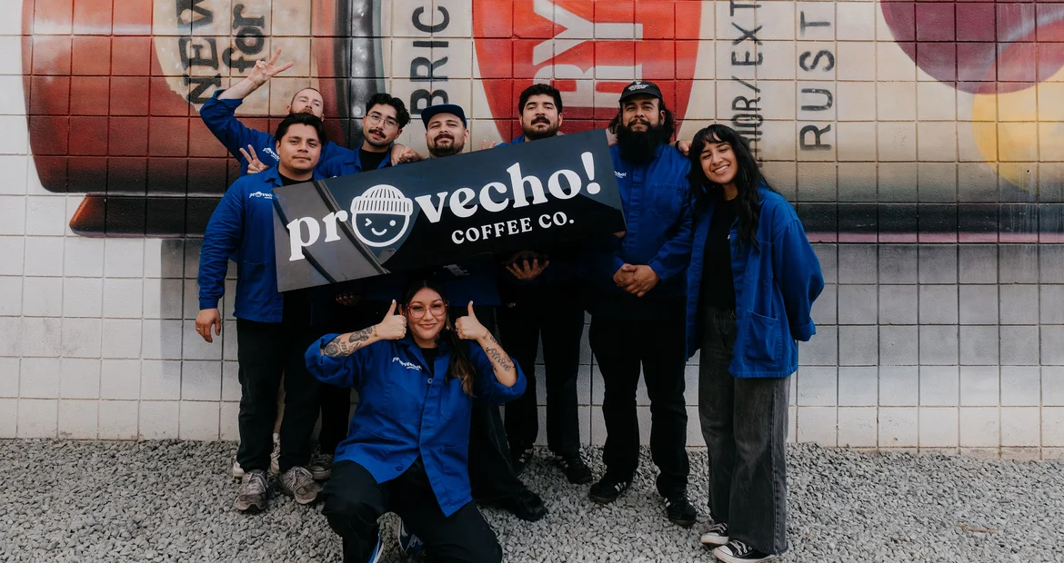 The Provecho team in Provecho workwear holding a Provecho! Coffee Co. banner in front of a painted mural