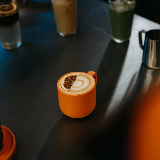 A cappuccino with latte art in an orange cup on the bar, an iced latte and an iced matcha behind it